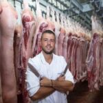 en una de las mejores carnicerías, un carnicero posa con cuchillos en las manos, en un cuarto lleno de carne de cerdo