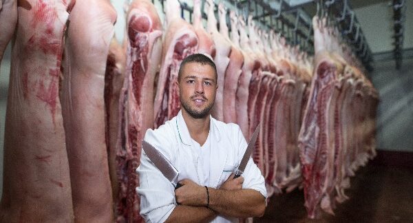 en una de las mejores carnicerías, un carnicero posa con cuchillos en las manos, en un cuarto lleno de carne de cerdo