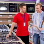 en las mejores tiendas de electrodomésticos, un encargado con camisa roja le muestra una cocina a una pareja, en una tienda
